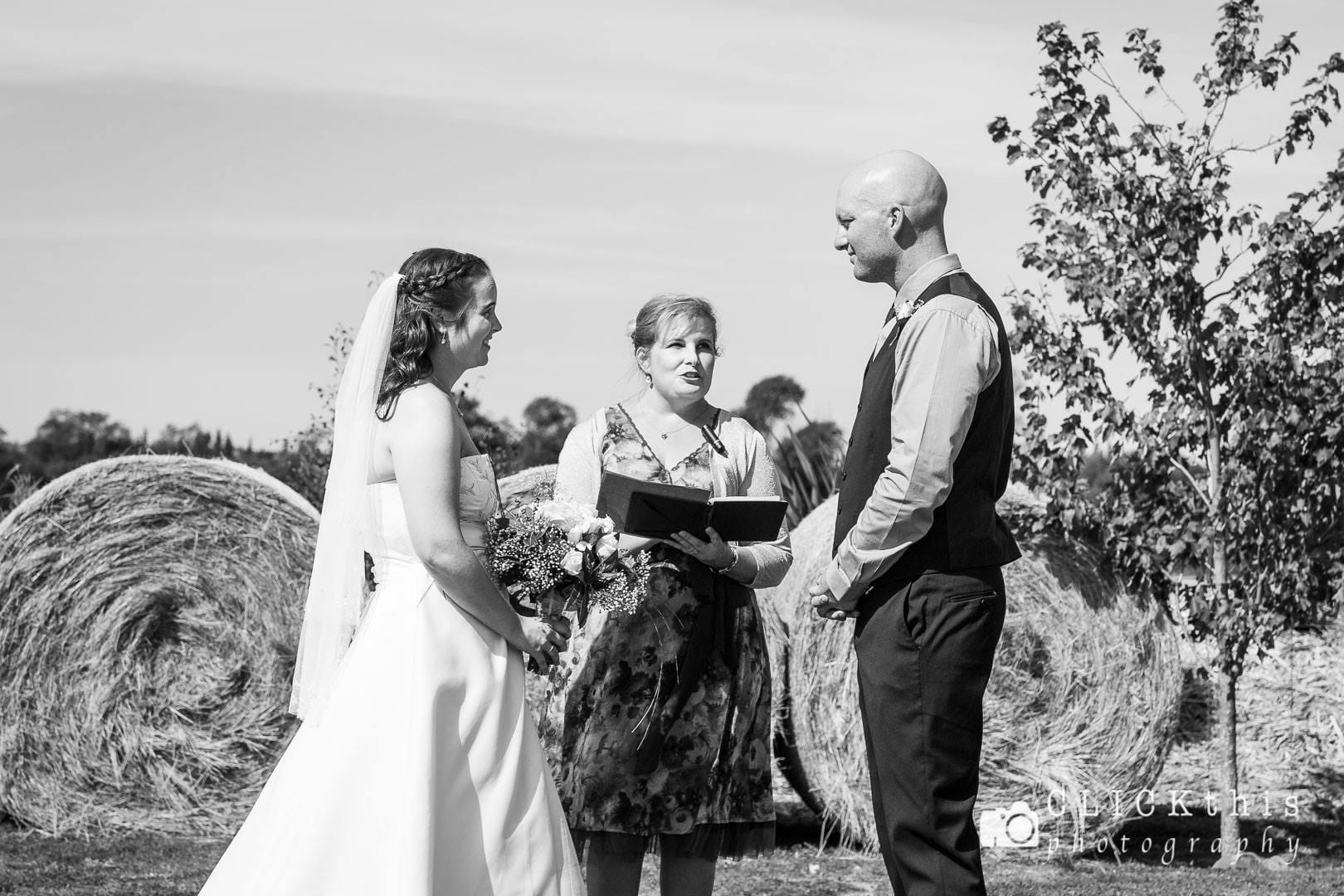 Black and white image of the bride and groom standing with Ana as she speaks during their wedding ceremony. Large round hay bales and a small tree are in the background.