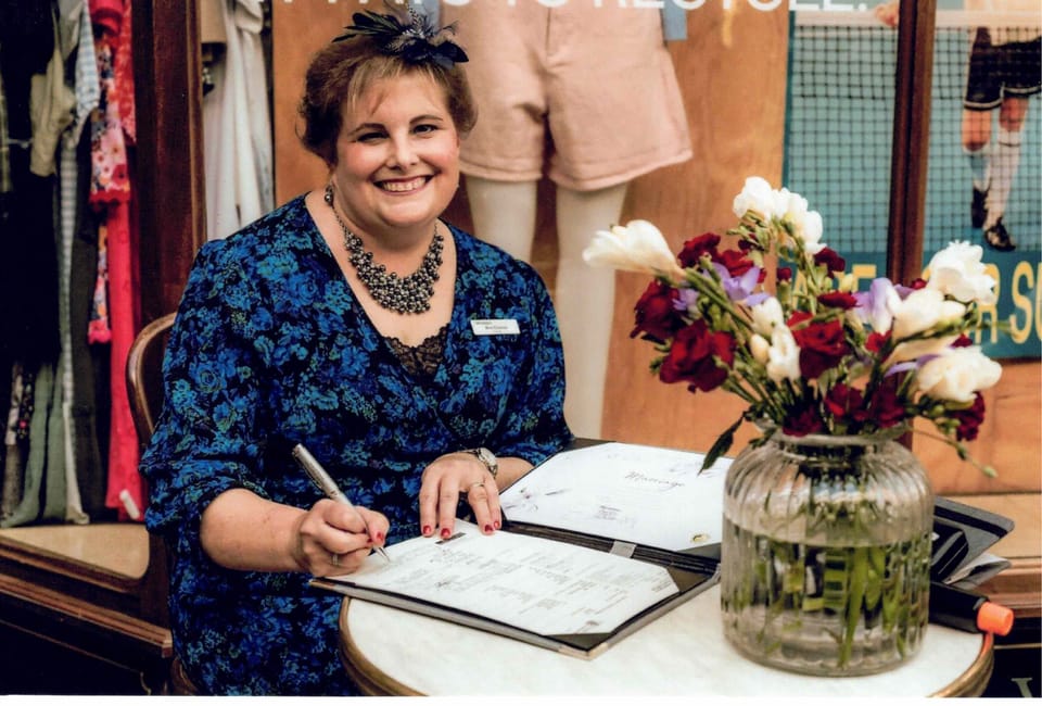Ana Connor, Celebrant is wearing a dark blue floral dress and matching hairpiece. She smiles as she signs the marriage registration papers at a wedding.